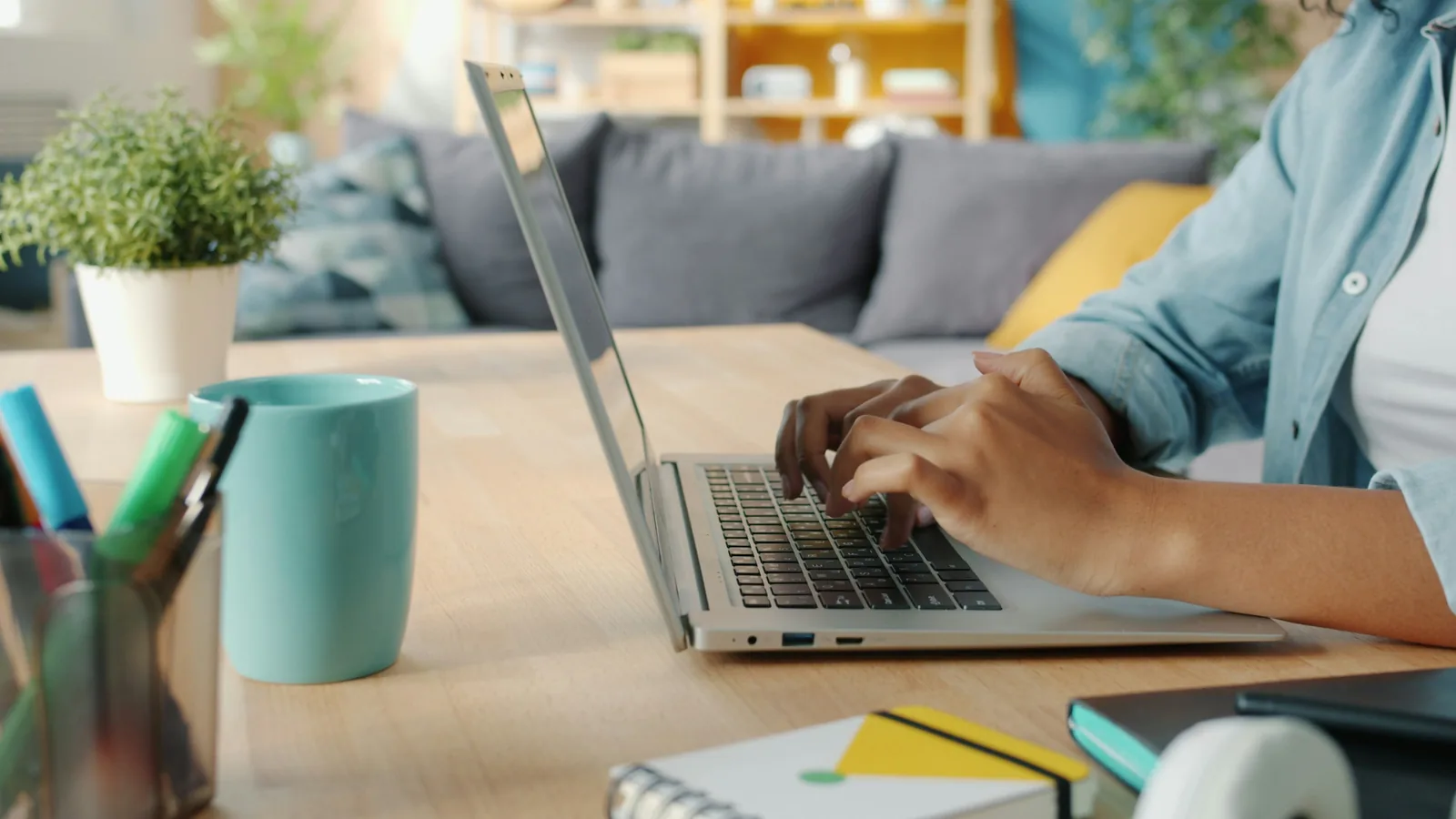 Hands typing on a laptop at a desk, next to a plant, a mug and a cup of pens