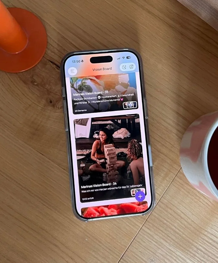My iPhone on a wooden table next to a candle and a cup of tea, showing two of my boards in the VISIYA app (app set to German): "Marinas Vision Board – 35" and "Marinas Vision Board – 34", marked 12/22 fulfilled