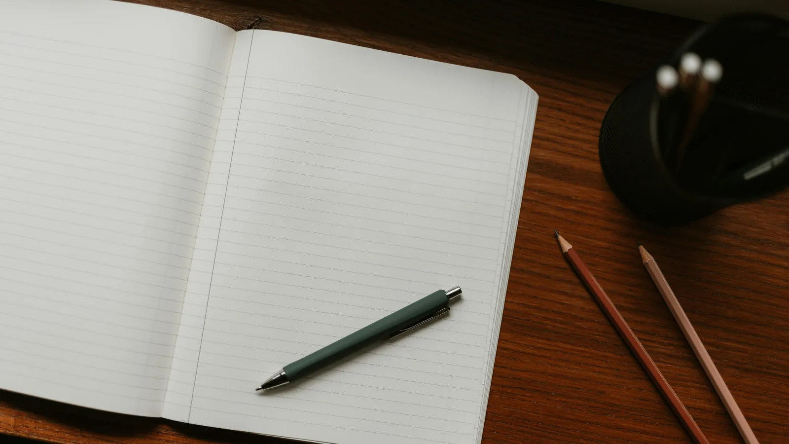 An open, blank notebook with a pen and two pencils on a wooden desk