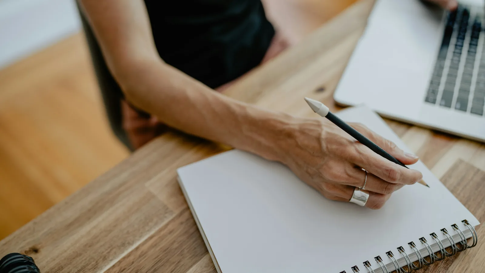A hand holding a pencil over a blank notebook, a laptop beside it on a wooden desk