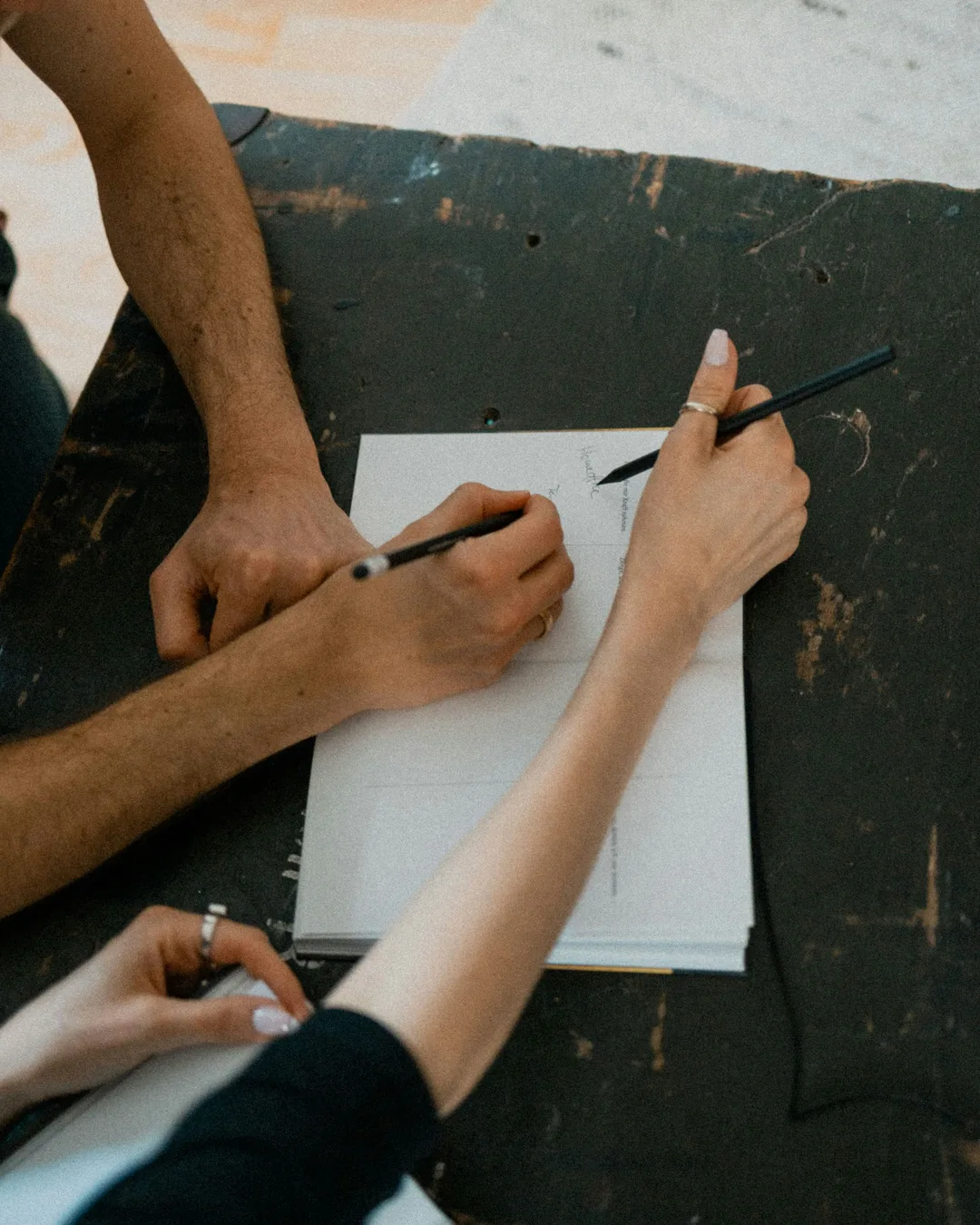 Two people's hands working on the same stack of paper on a table, seen from above