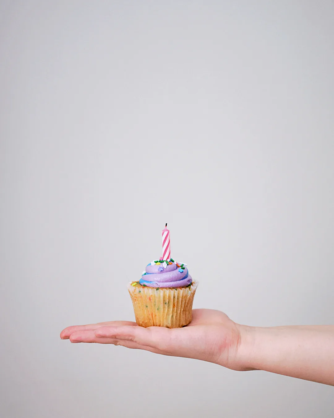 A hand holding a cupcake with a single lit candle against a pale gray background