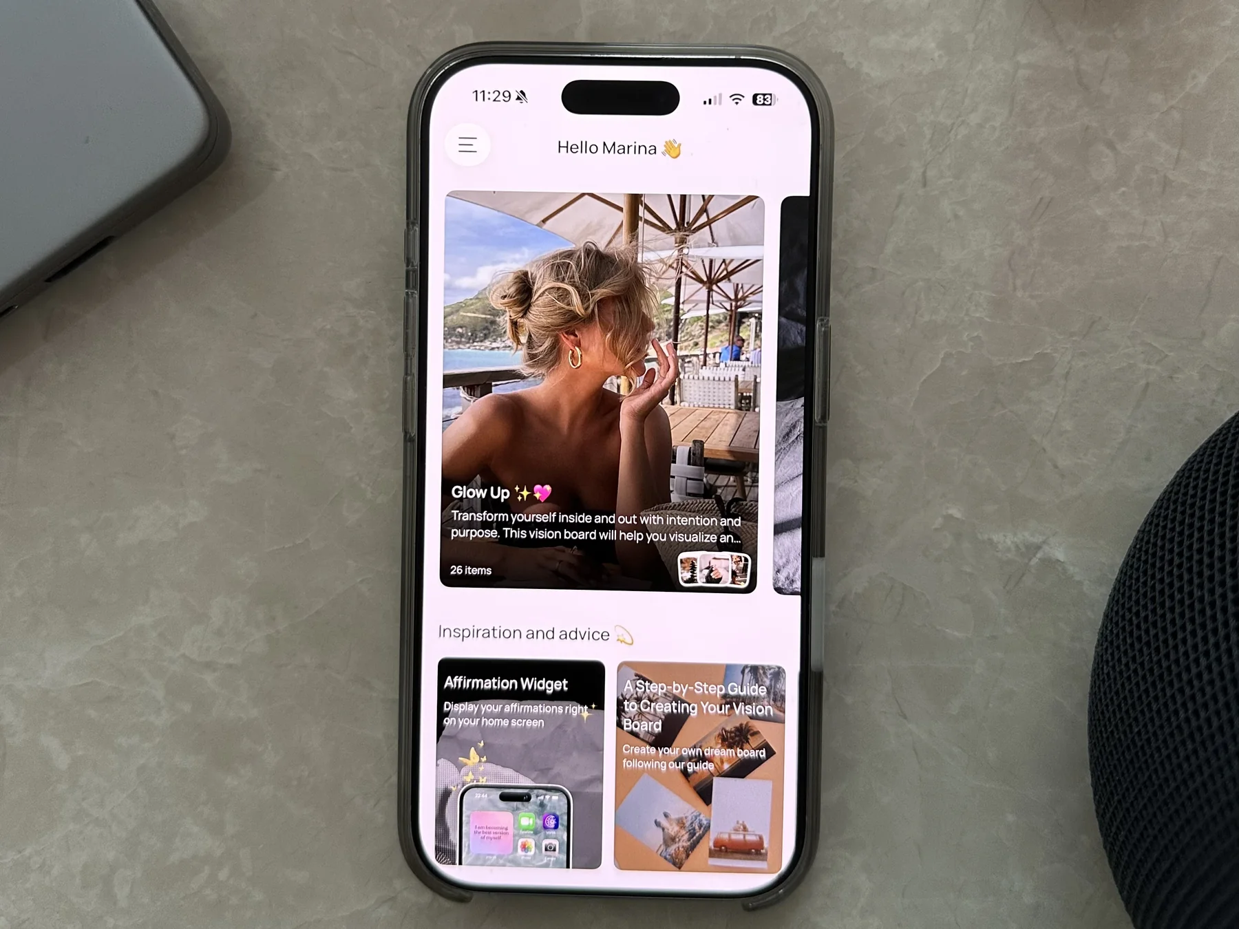 An iPhone on a marble table showing the VISIYA app with the Glow Up board and affirmations
