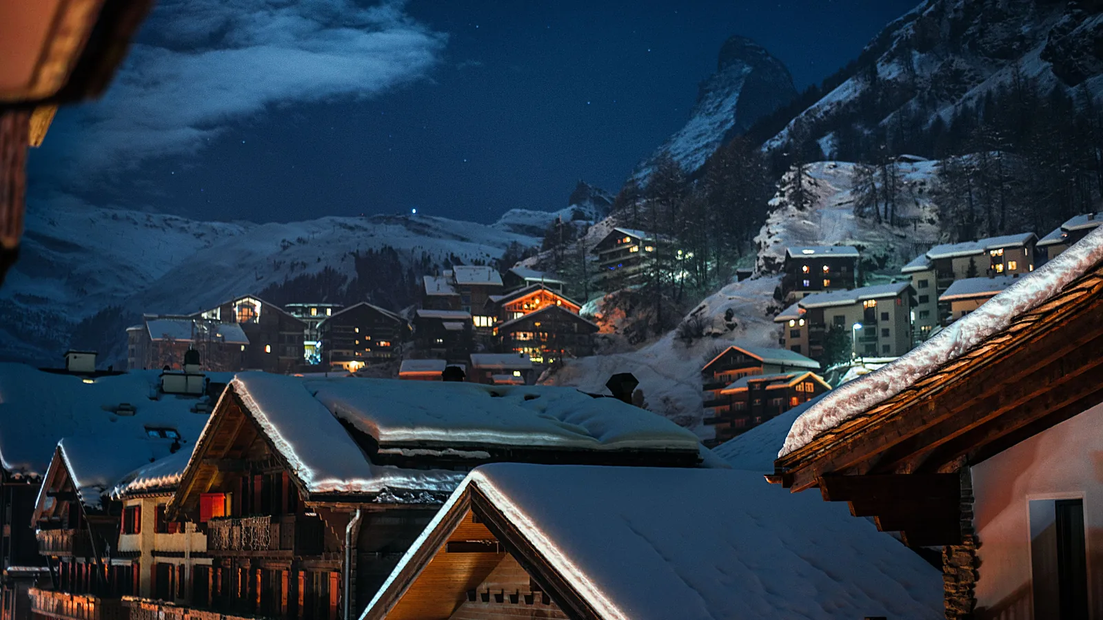 Snow-covered wooden chalets in an Alpine village at night, mountains behind