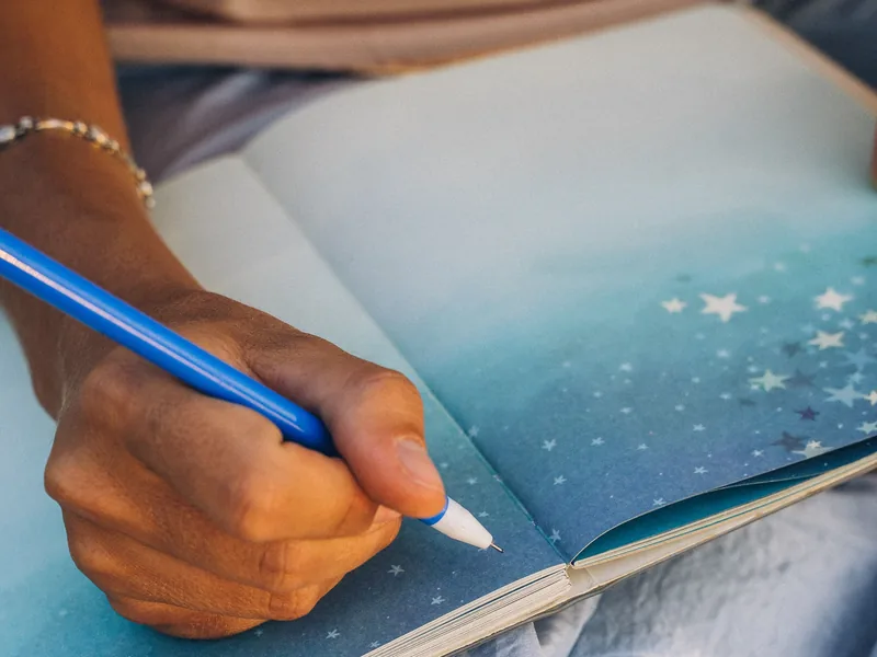 A hand writing with a blue pencil in a notebook with a starry night sky on its pages