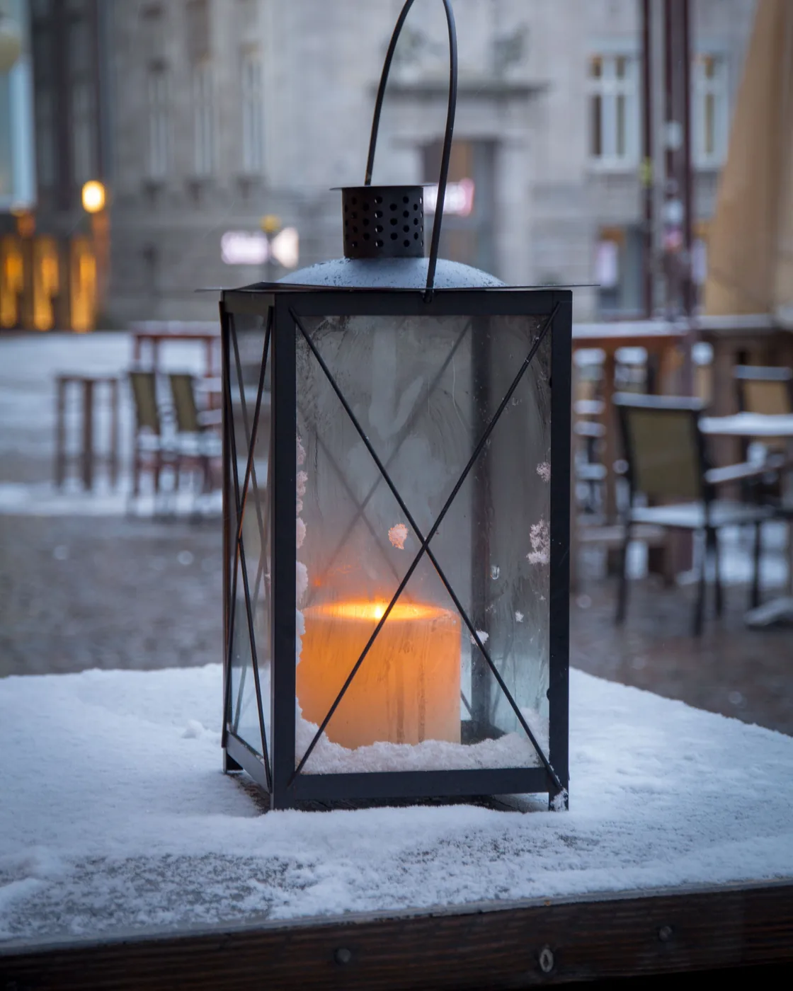 A candle burning inside a lantern on a snowy table outside a quiet café