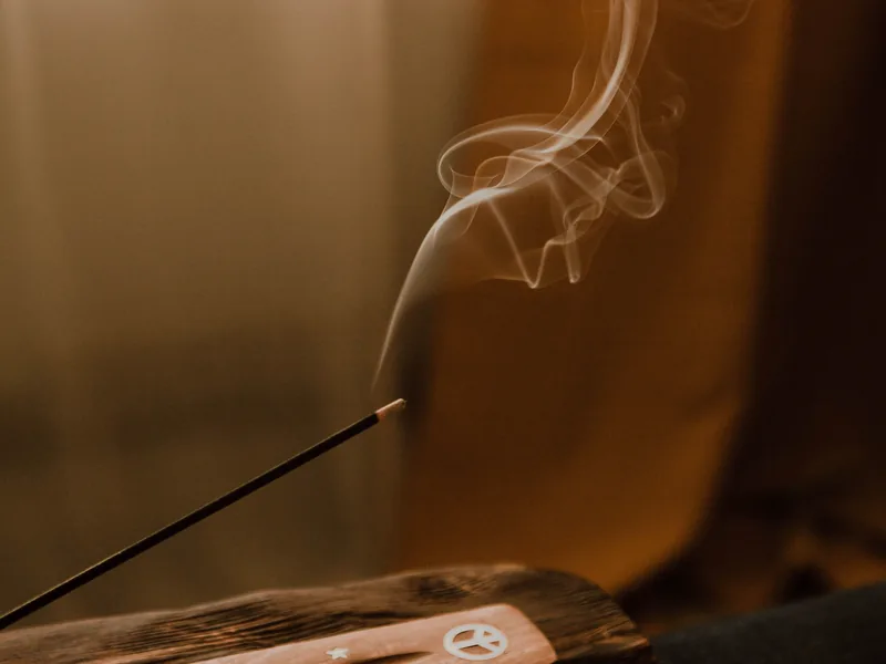 Smoke curling up from an incense stick in a wooden holder by a window