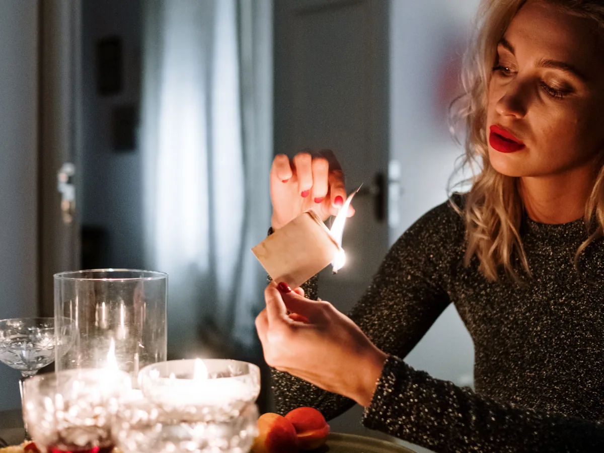 A woman at a candlelit dinner table setting a folded slip of paper alight with a match