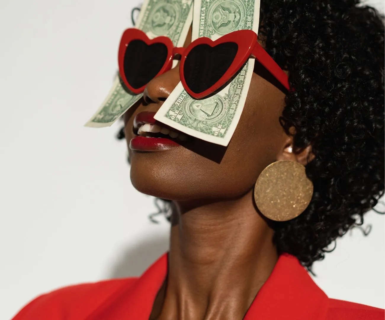 Eine lächelnde Frau im roten Anzug mit herzförmiger Sonnenbrille und Dollarscheinen als Symbol für ein positives Geld-Mindset