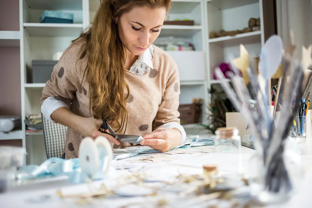 Frau an einem Basteltisch schneidet mit der Schere Papierbilder für ein selbstgemachtes Vision Board aus