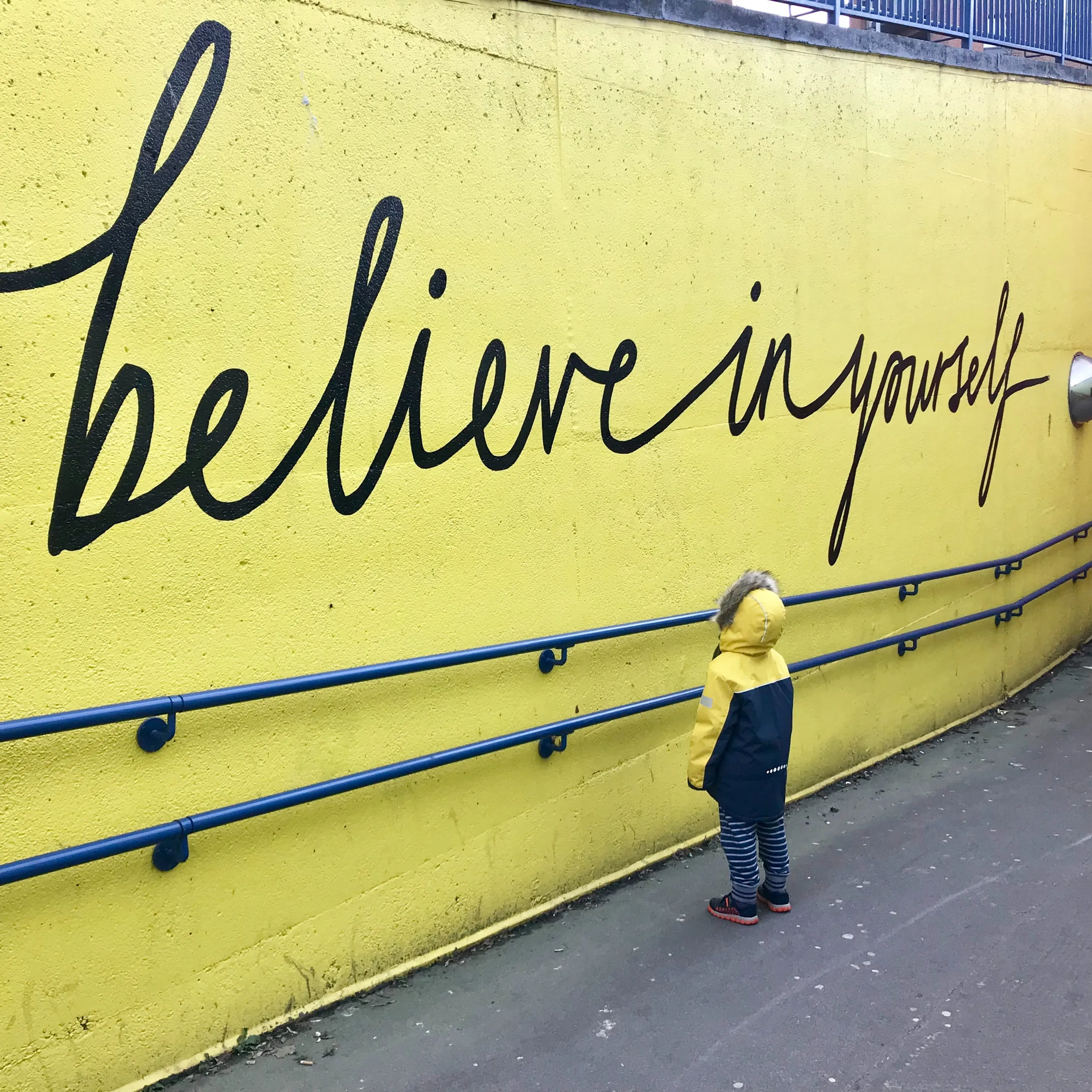 A person writing the words 'Believe in yourself' on a bright yellow wall