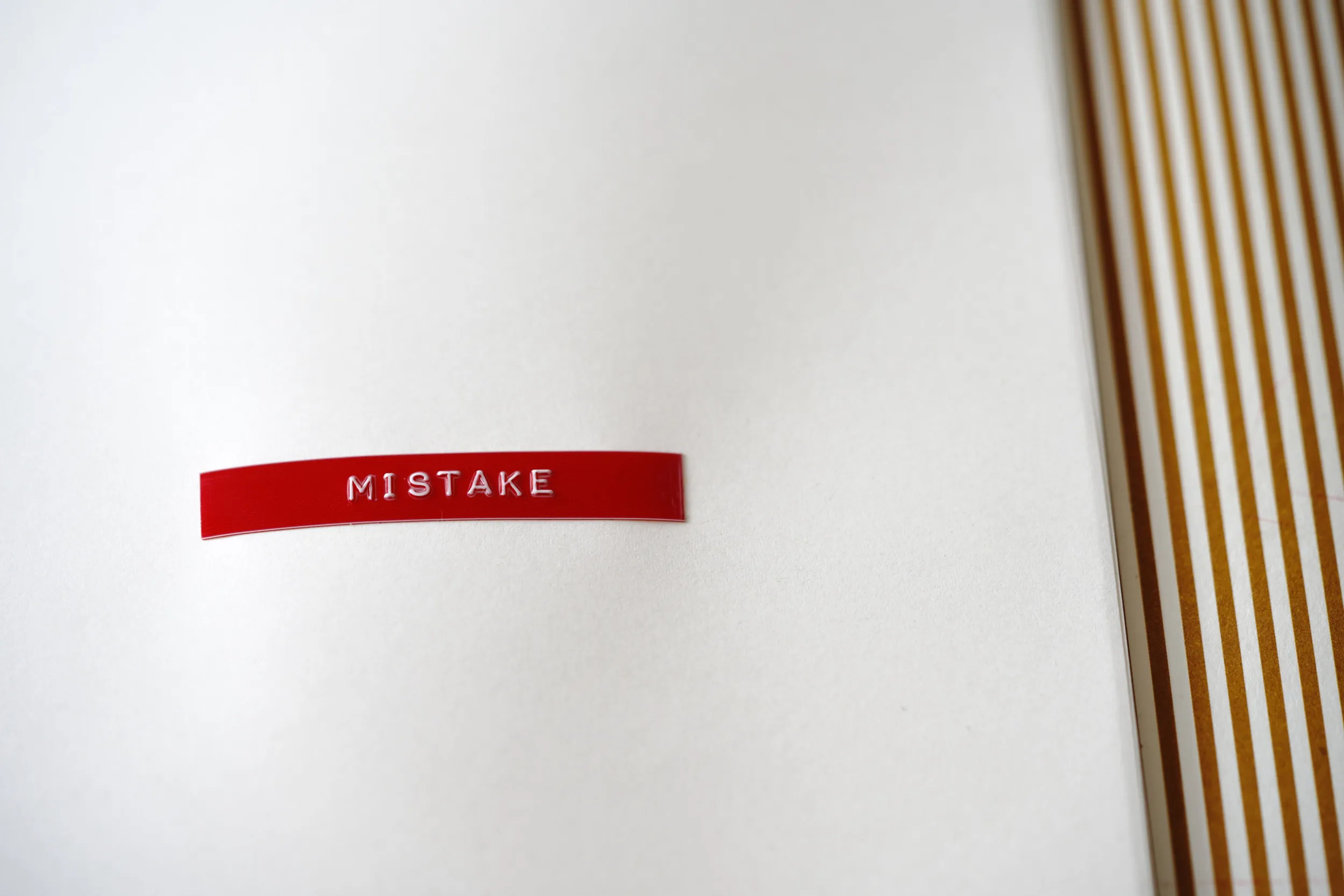 Red embossed label reading 'MISTAKE' on a cream page beside soft gold stripes