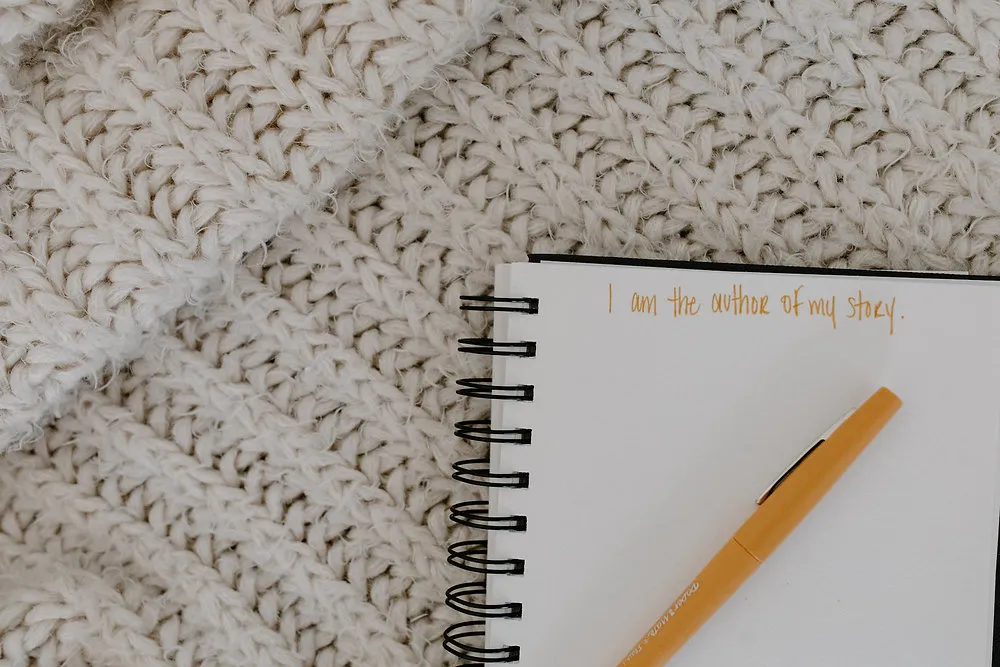Aufgeschlagenes Spiralnotizbuch auf kuscheliger Strickdecke, goldener Stift und der handgeschriebene Satz 'I am the author of my story'