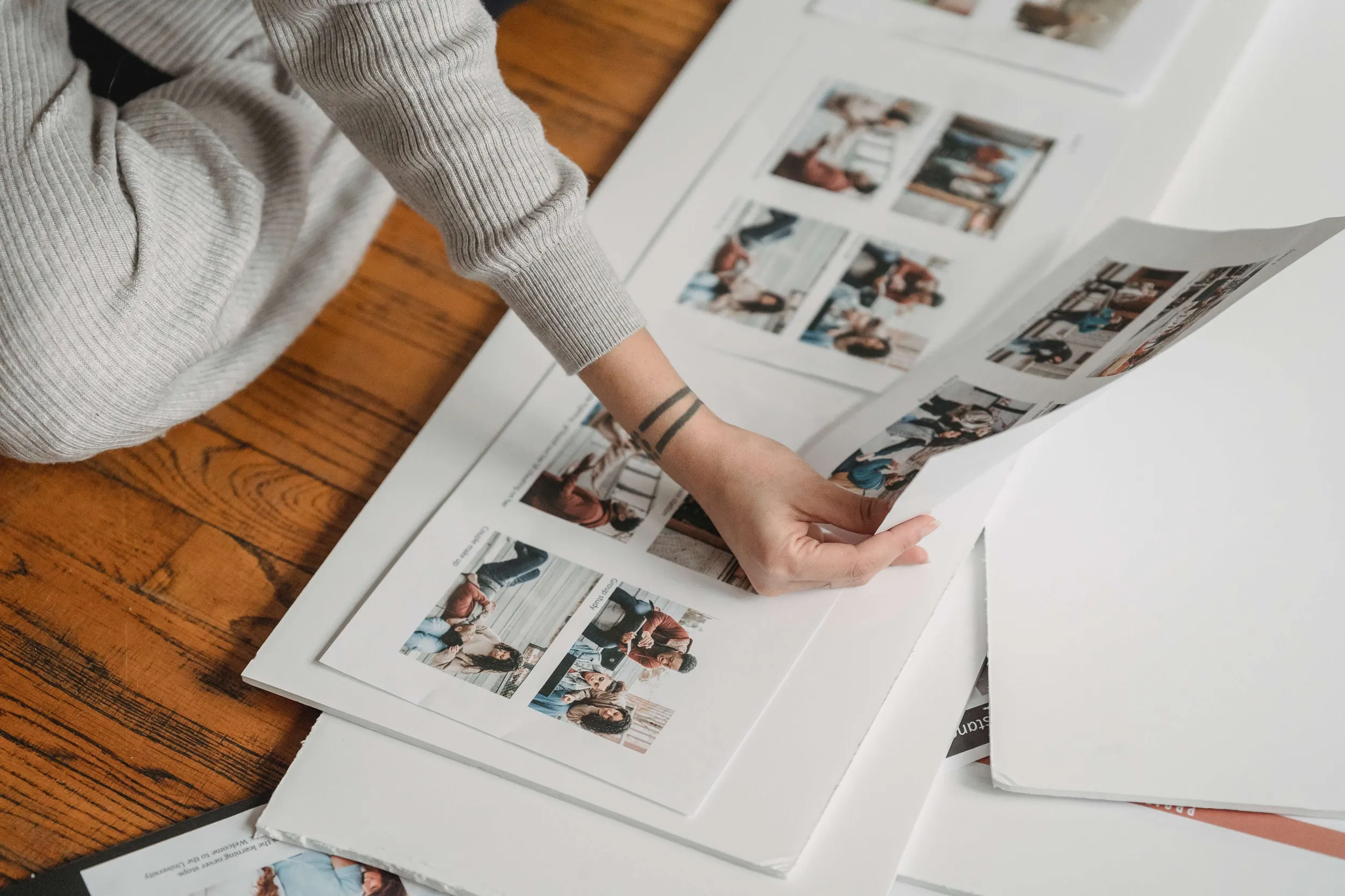 Person in a grey sweater arranging printed photo sheets on a wooden floor to pick vision board images
