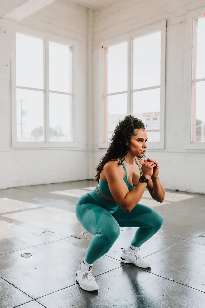 Woman in teal activewear doing a deep squat in a bright, sunlit studio