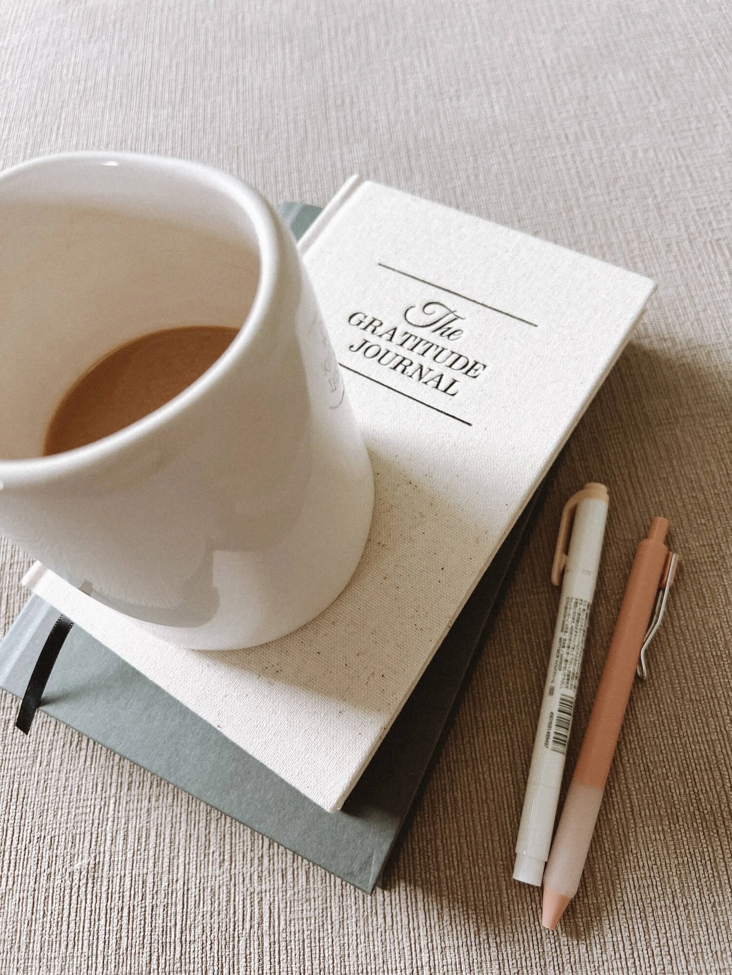 Cream mug of coffee resting on a book titled 'The Gratitude Journal' beside two pastel pens