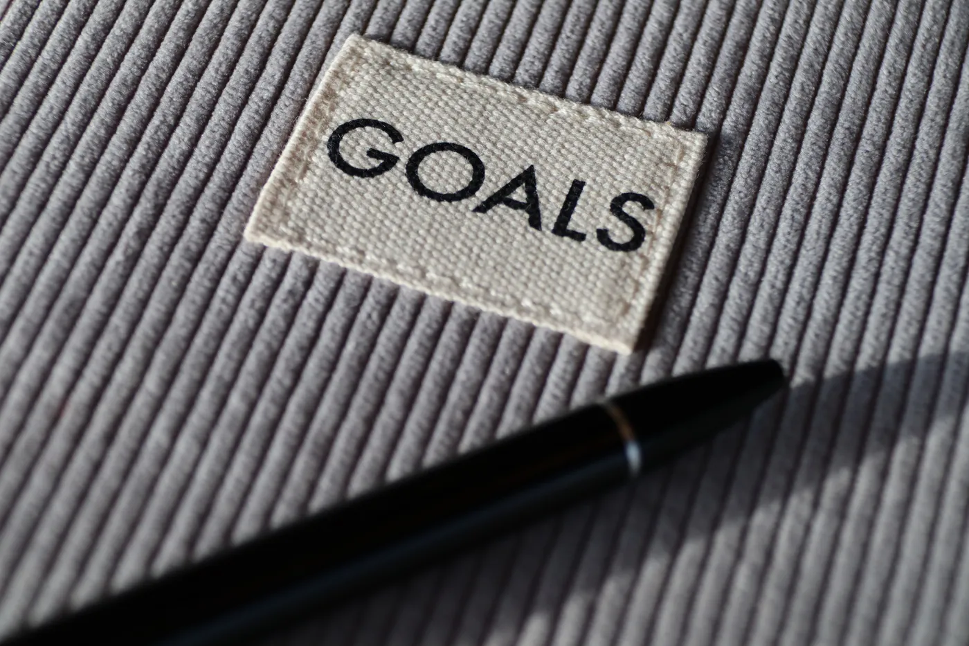 A fabric label reading 'GOALS' on grey ribbed cloth with a sleek black pen resting beside it