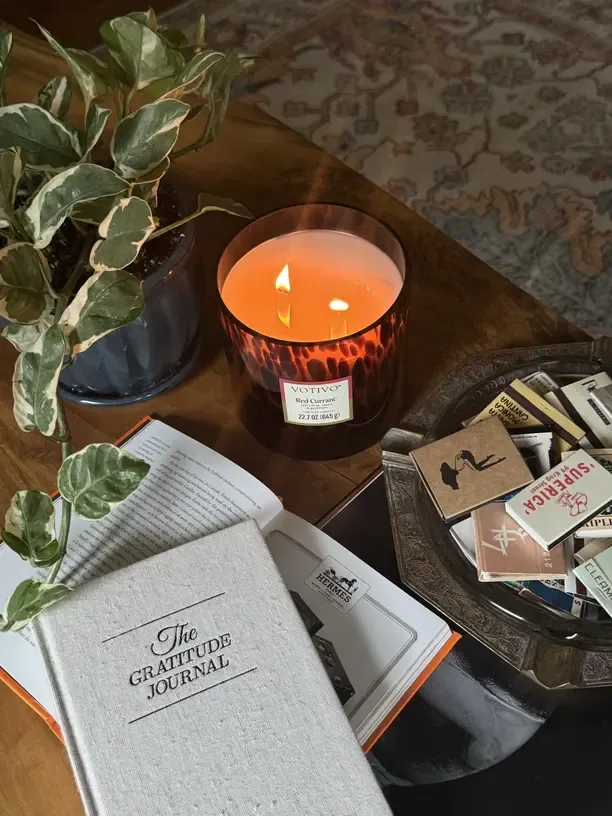 Linen 'The Gratitude Journal' beside a lit tortoiseshell candle and a trailing plant on a cozy dark table