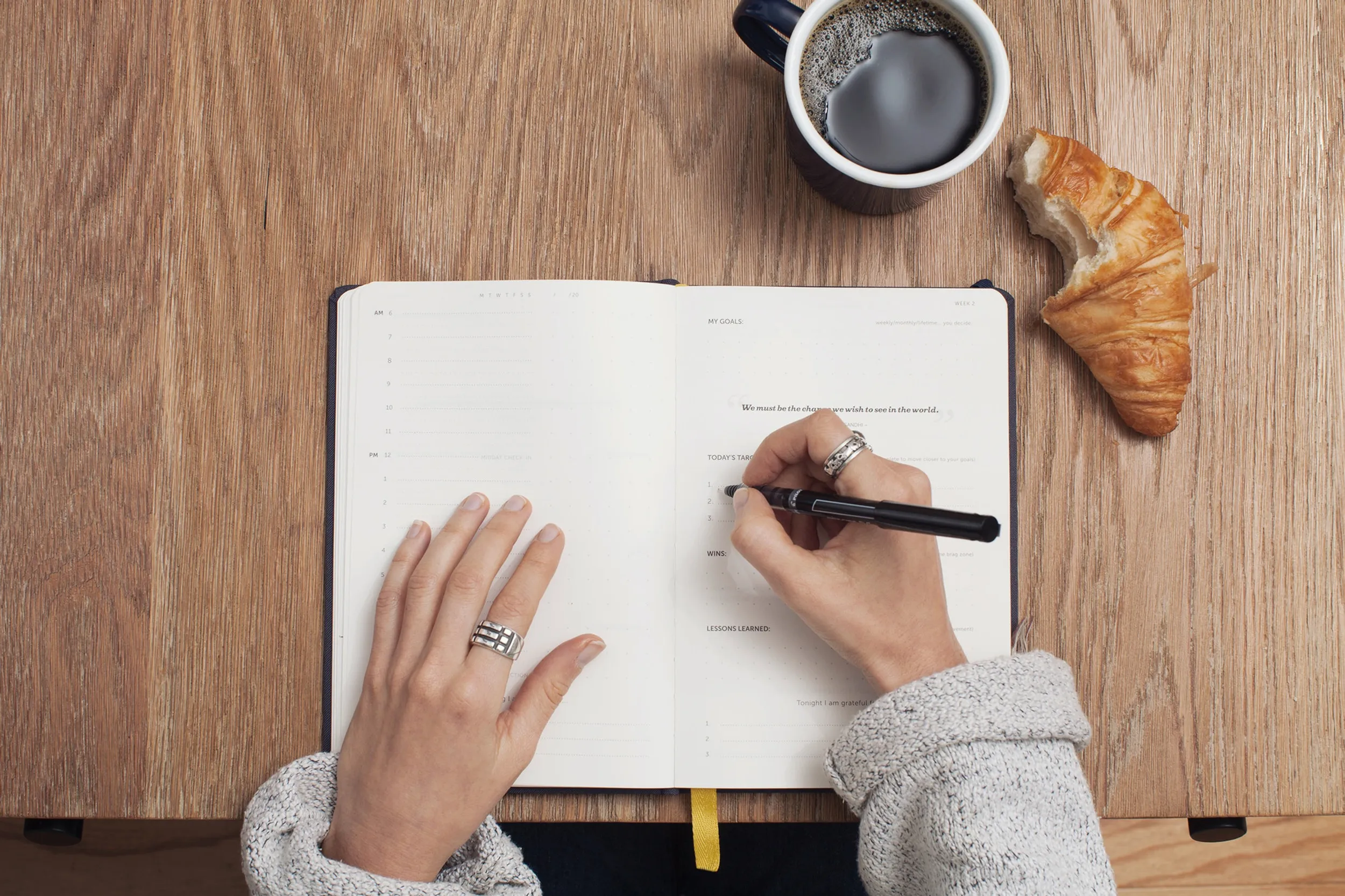Overhead view of hands writing goals in an open planner beside black coffee and a croissant