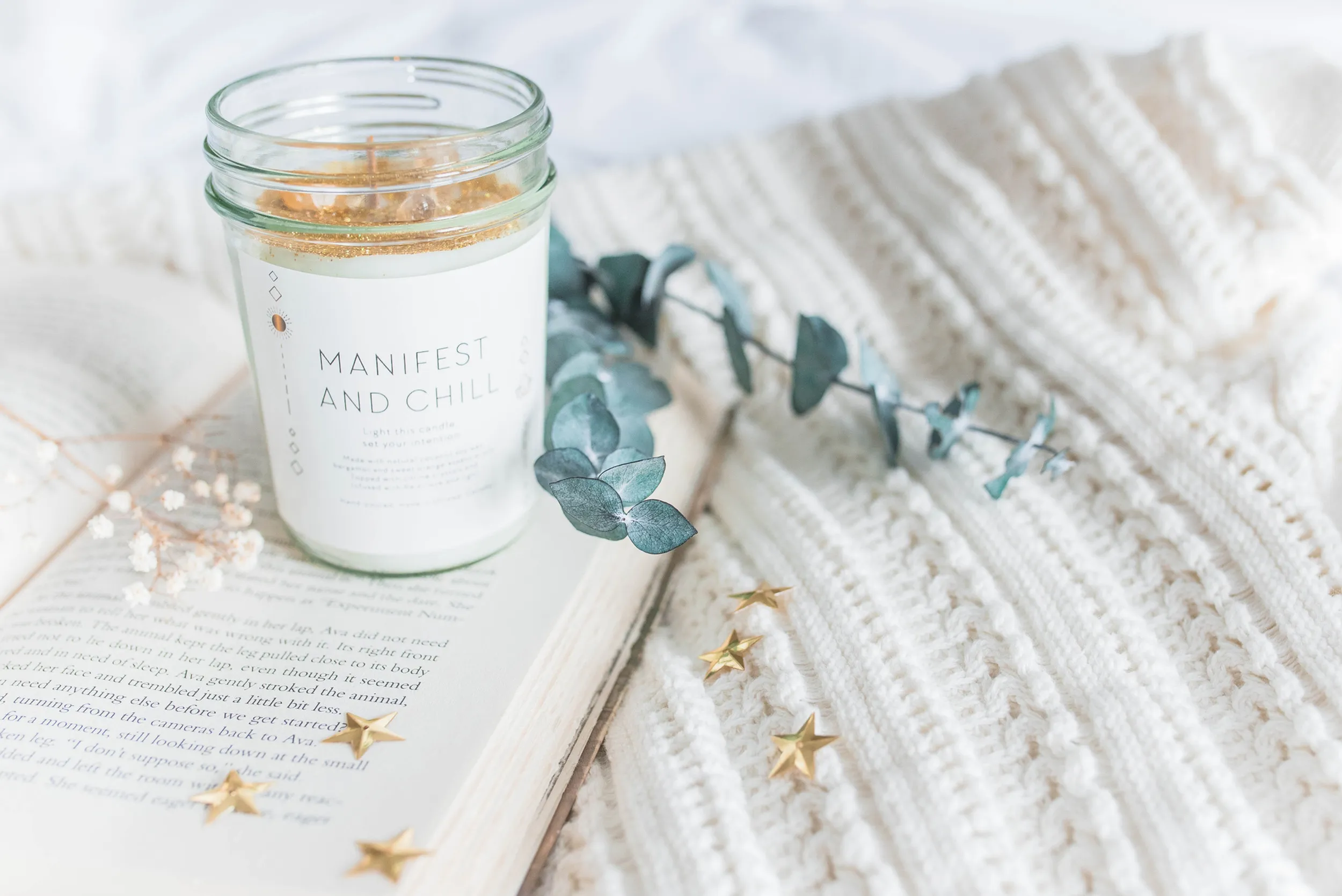 Candle labelled Manifest and Chill resting on an open book with eucalyptus, gold stars and a knit throw