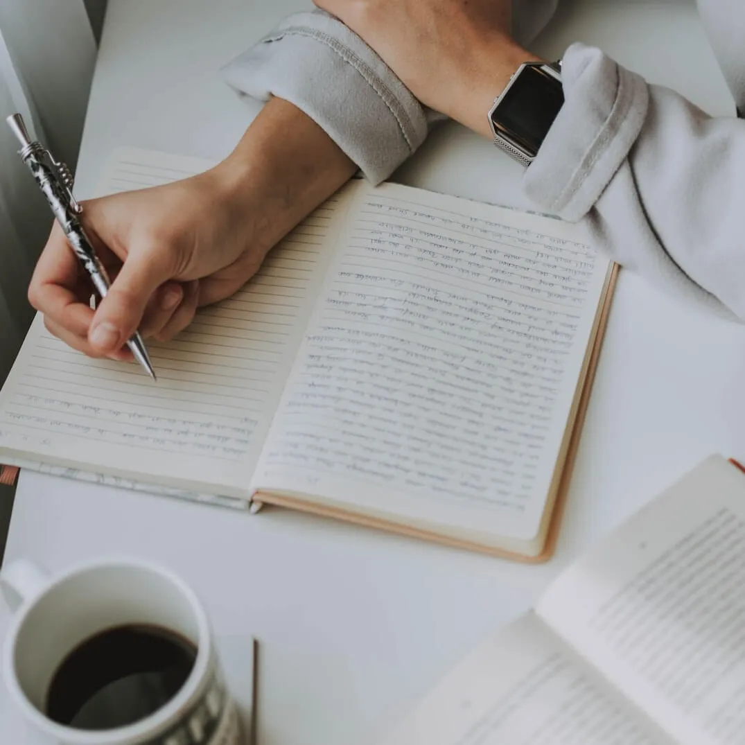 Overhead view of a hand writing in a lined journal full of cursive, a smartwatch on the wrist and black coffee nearby