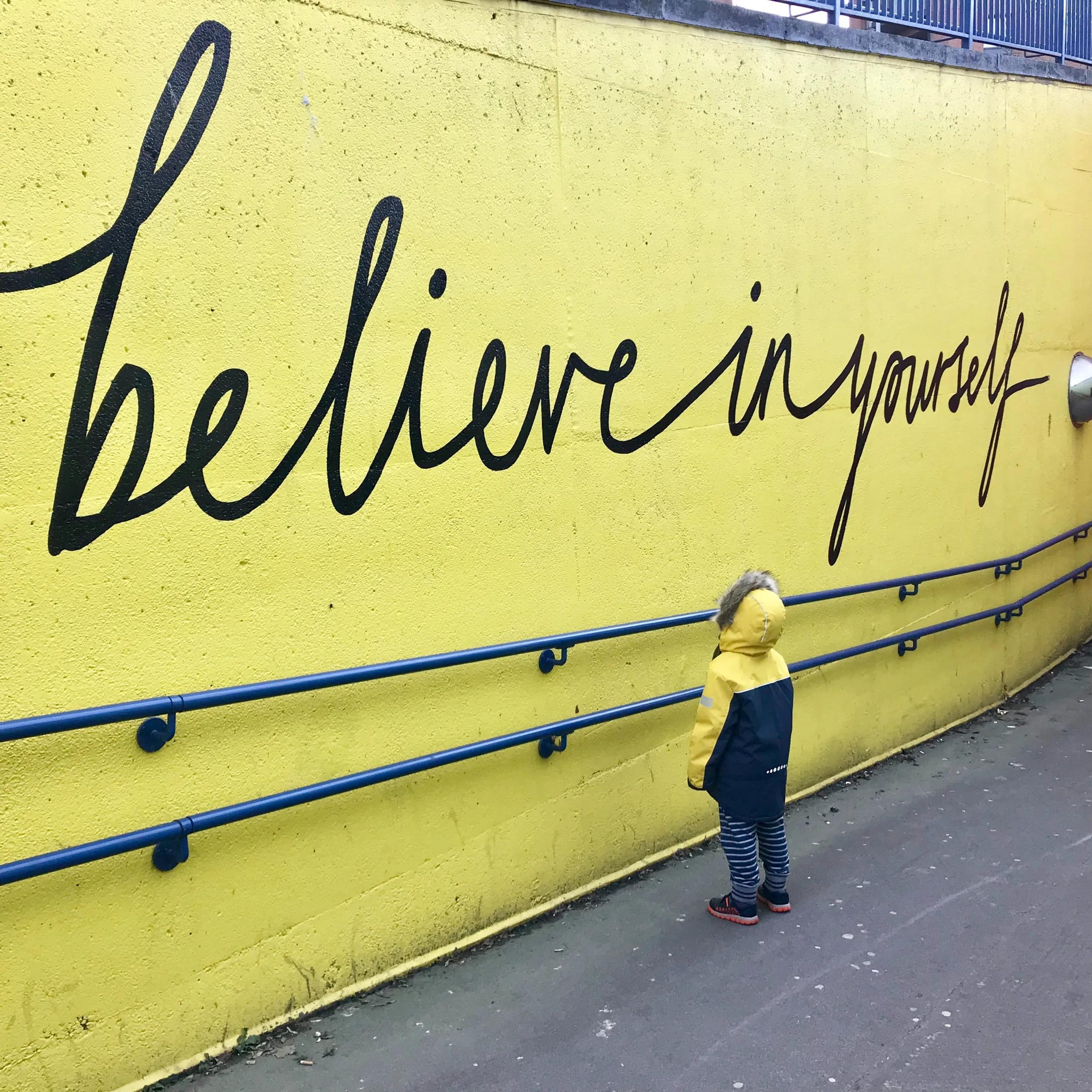 A person writing the words 'Believe in yourself' on a bright yellow wall