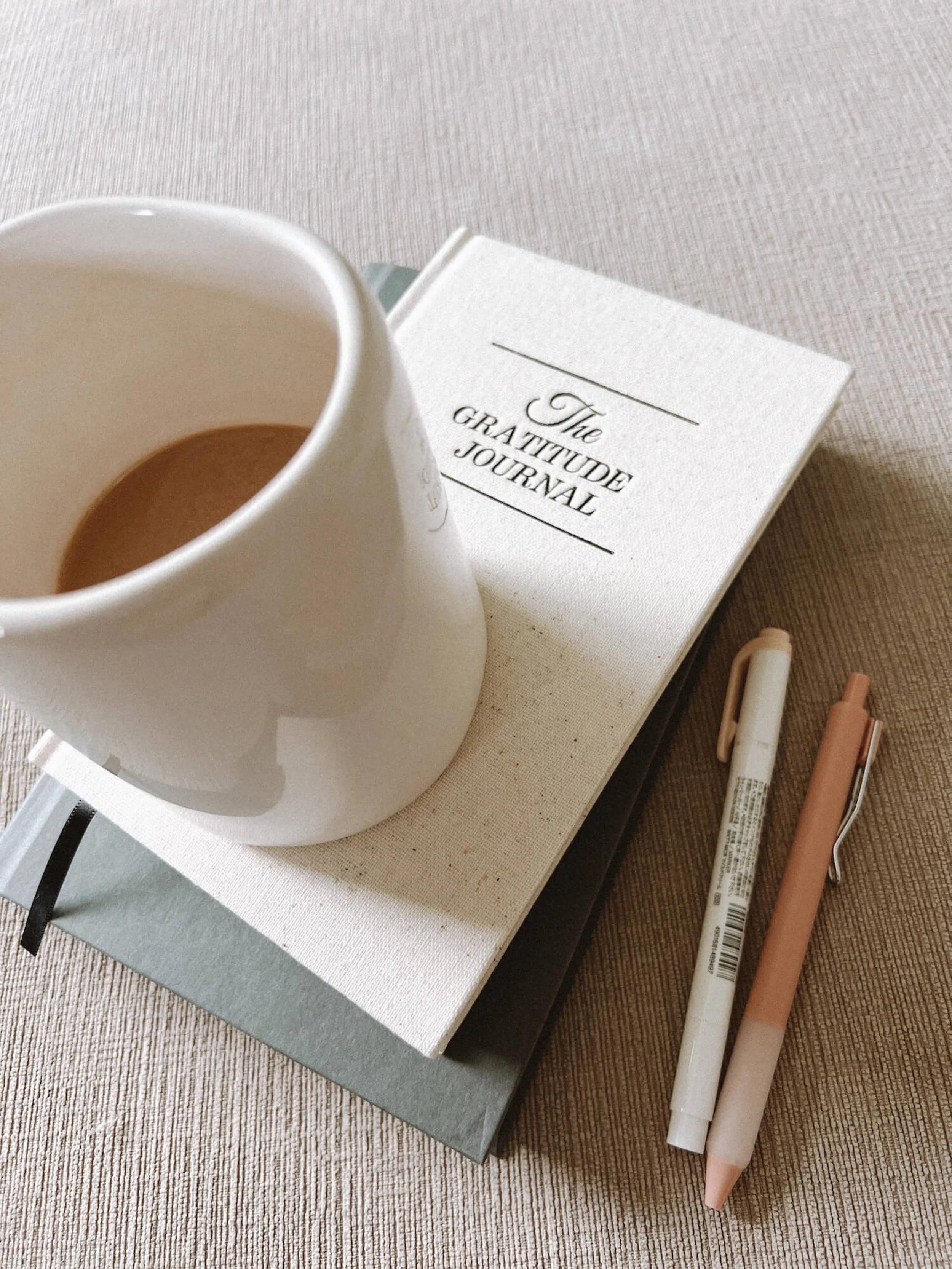 Cremefarbene Tasse Kaffee auf einem Buch mit dem Titel 'The Gratitude Journal' neben zwei Stiften