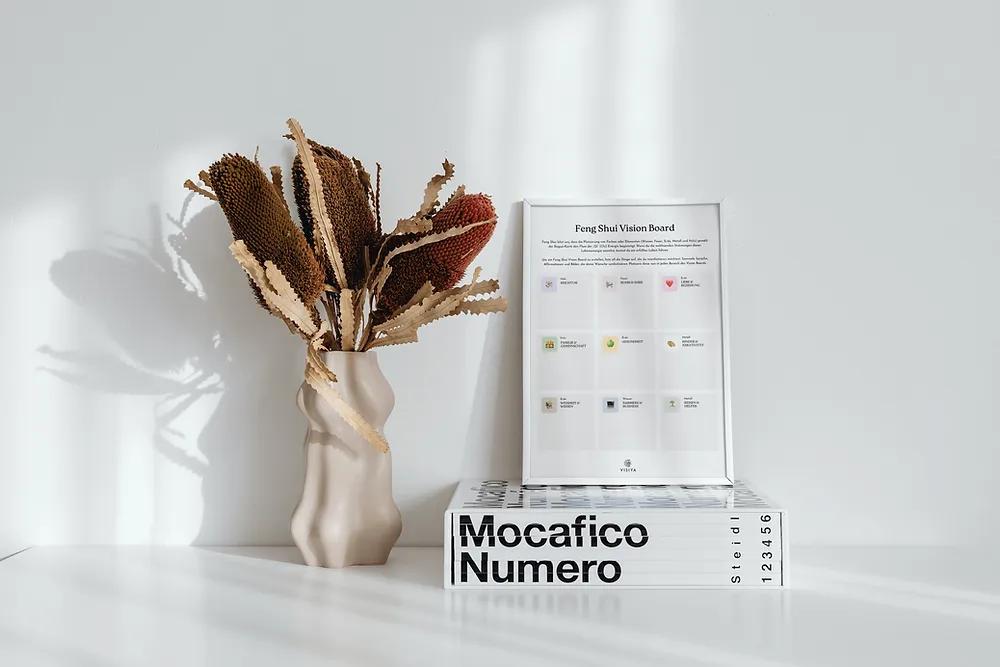 Gerahmte Feng Shui Vision Board Vorlage auf einem Bildband neben einer Vase mit Trockenblumen