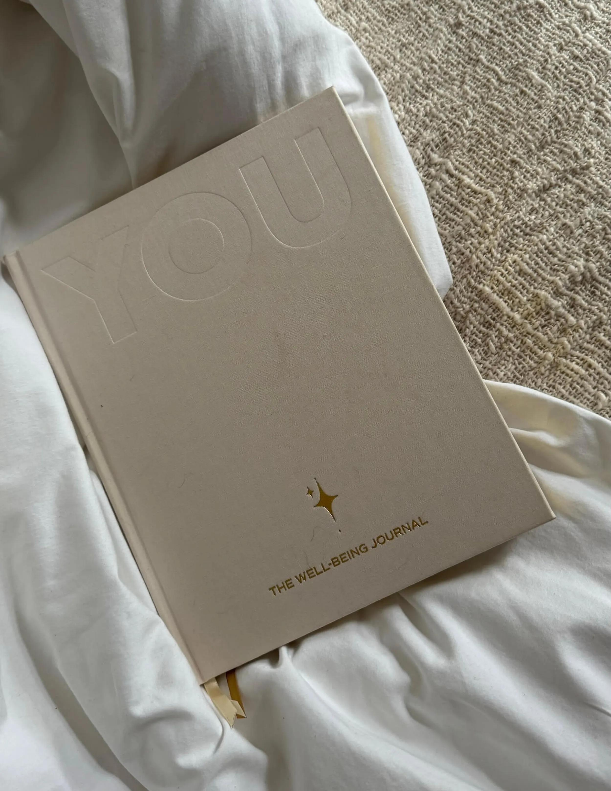 Cremefarbenes Leinen-Journal mit Prägung 'YOU' und goldenem Schriftzug 'The Well-Being Journal' auf weißer Bettwäsche
