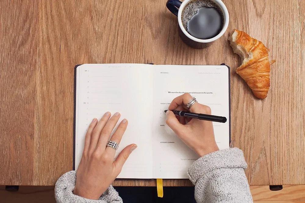 Hände schreiben Ziele in ein aufgeschlagenes Journal, daneben schwarzer Kaffee und ein Croissant