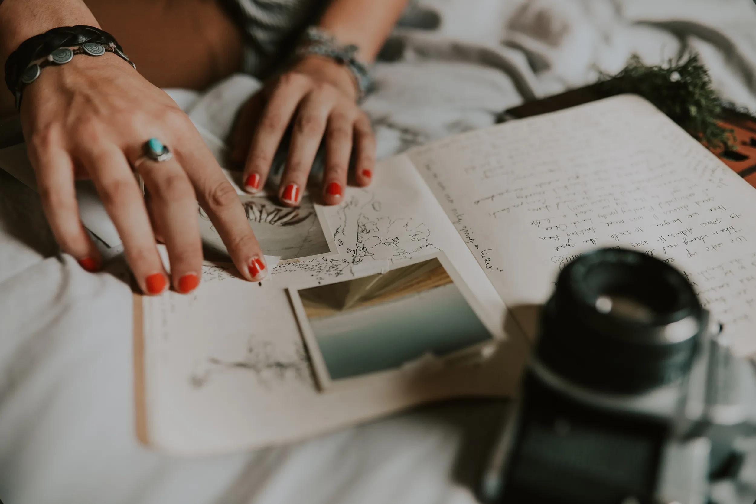 Hände mit rotem Nagellack ordnen Fotos in ein aufgeschlagenes Reisetagebuch, daneben eine analoge Kamera