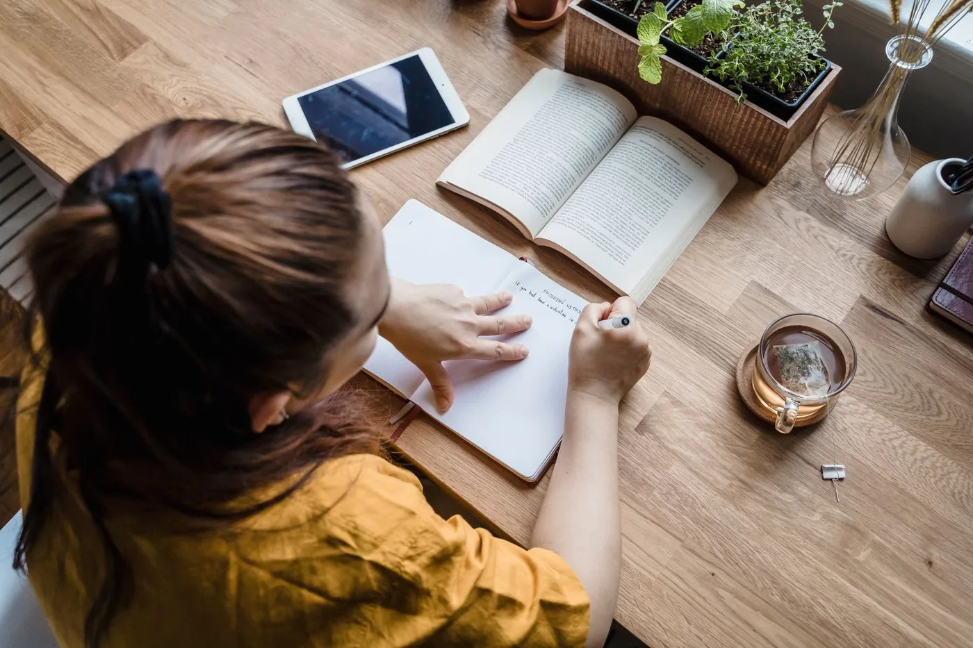 Женщина пишет в блокноте за деревянным столом с книгами, чаем и планшетом, оценивая баланс своей жизни