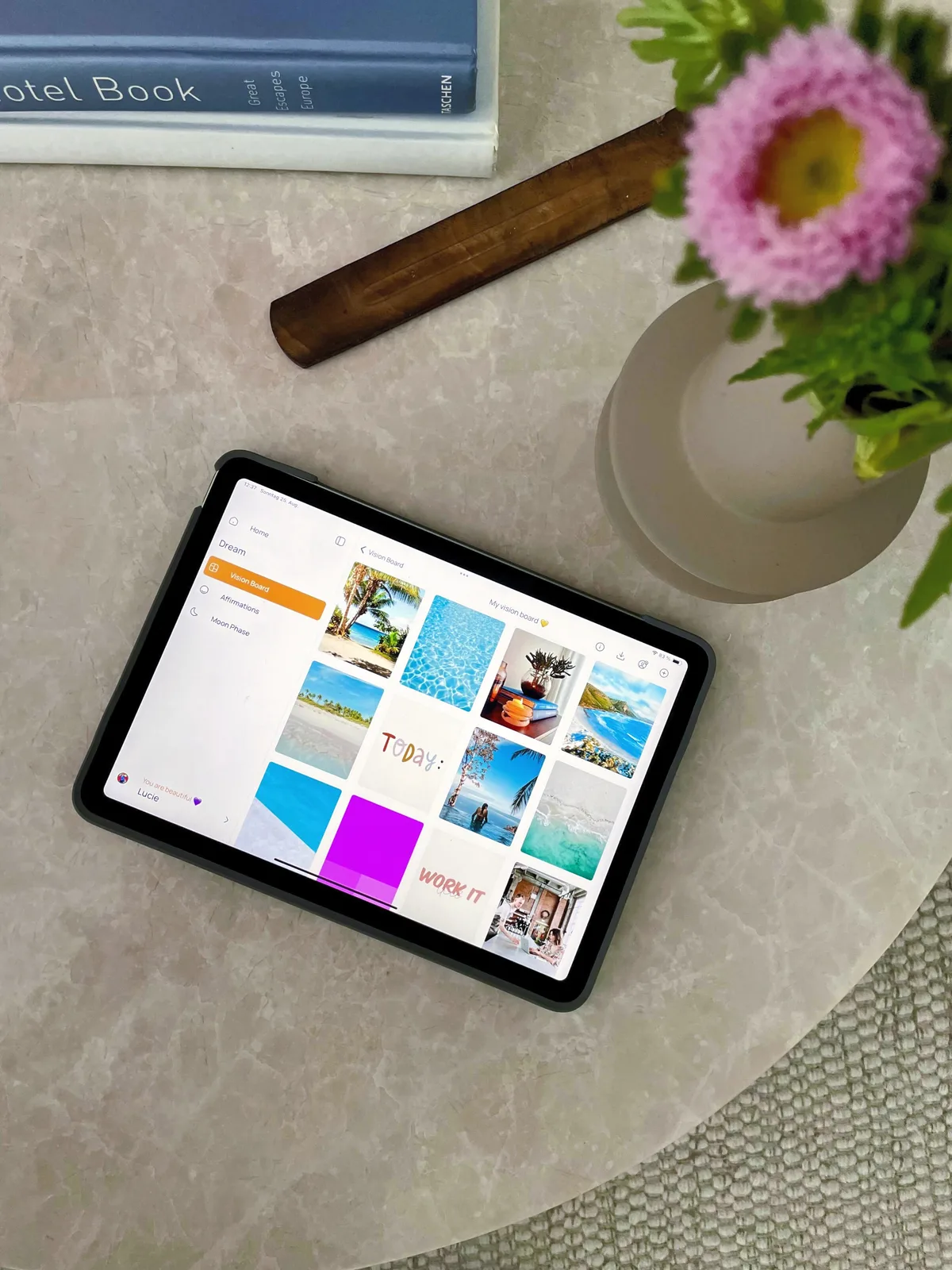 iPad с цифровой картой желаний из фотографий путешествий и пляжей в приложении VISIYA, на каменном столе рядом с книгой и вазой с цветами
