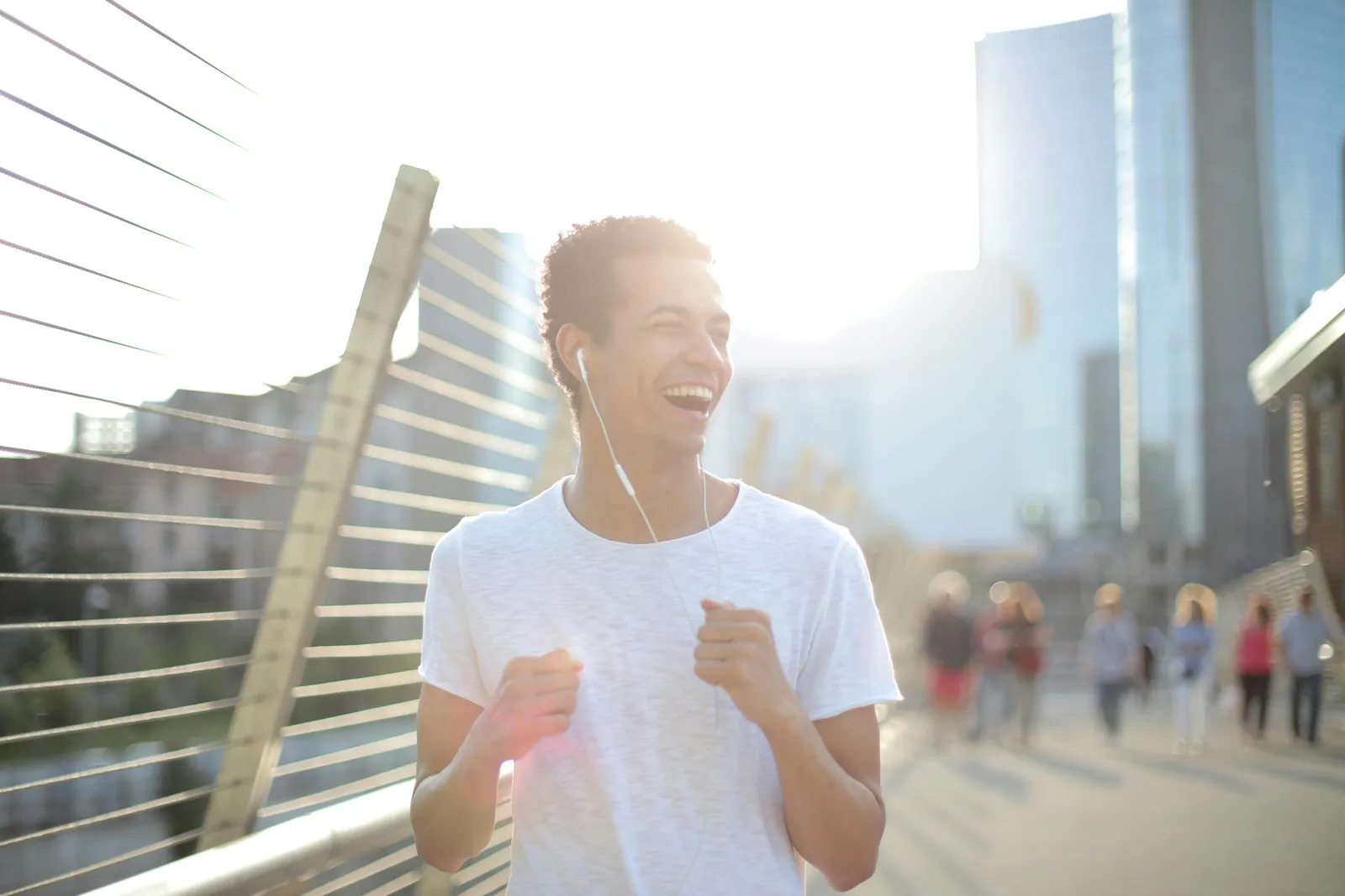 A smiling young man jogging with earphones on a sunlit city bridge