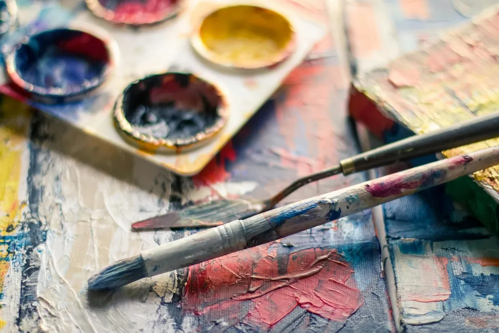 Watercolour paints and well-used brushes resting on a brightly painted canvas