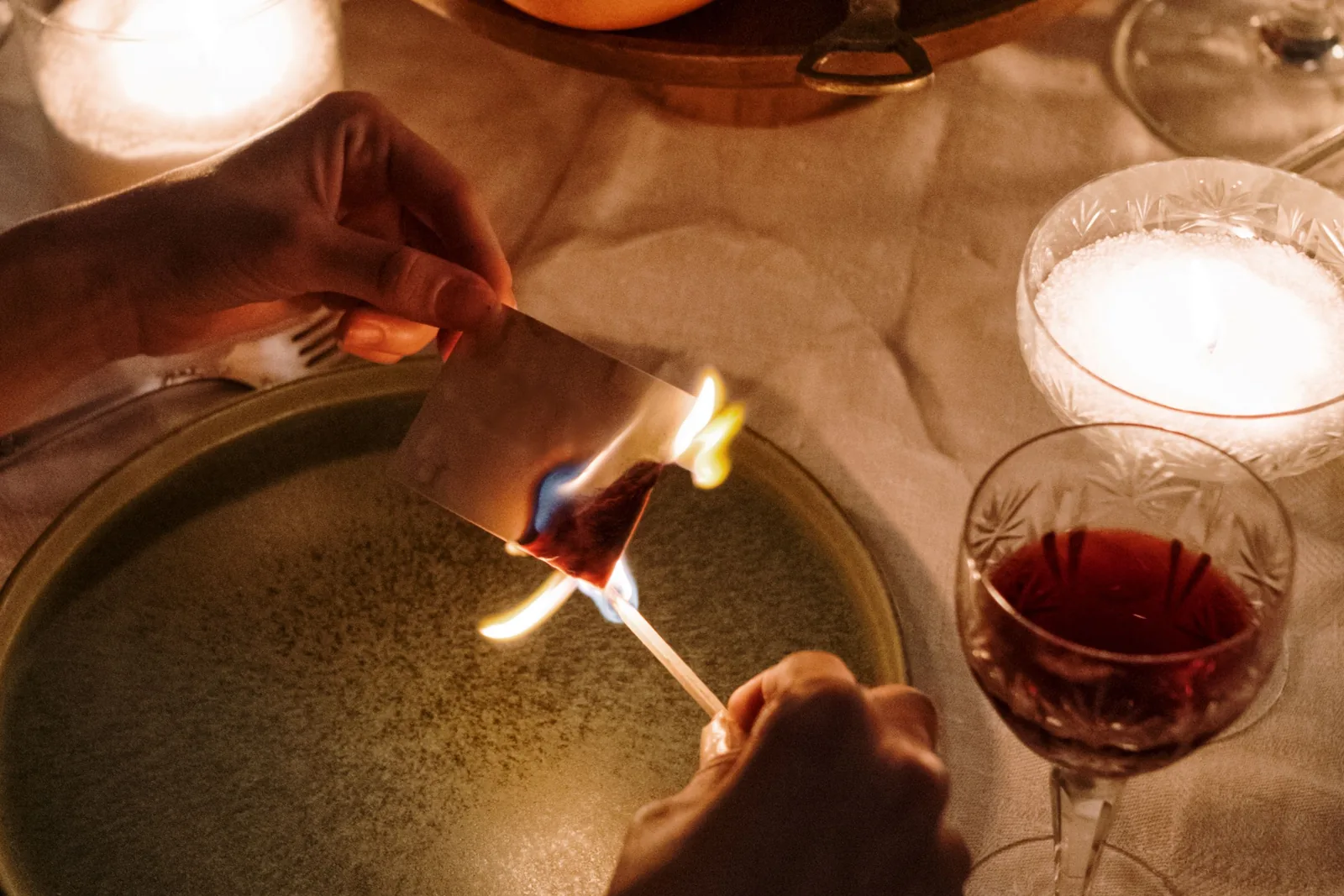 Hände zünden über einem Teller bei Kerzenlicht einen Wunschzettel mit einem Streichholz an, daneben ein Glas Rotwein